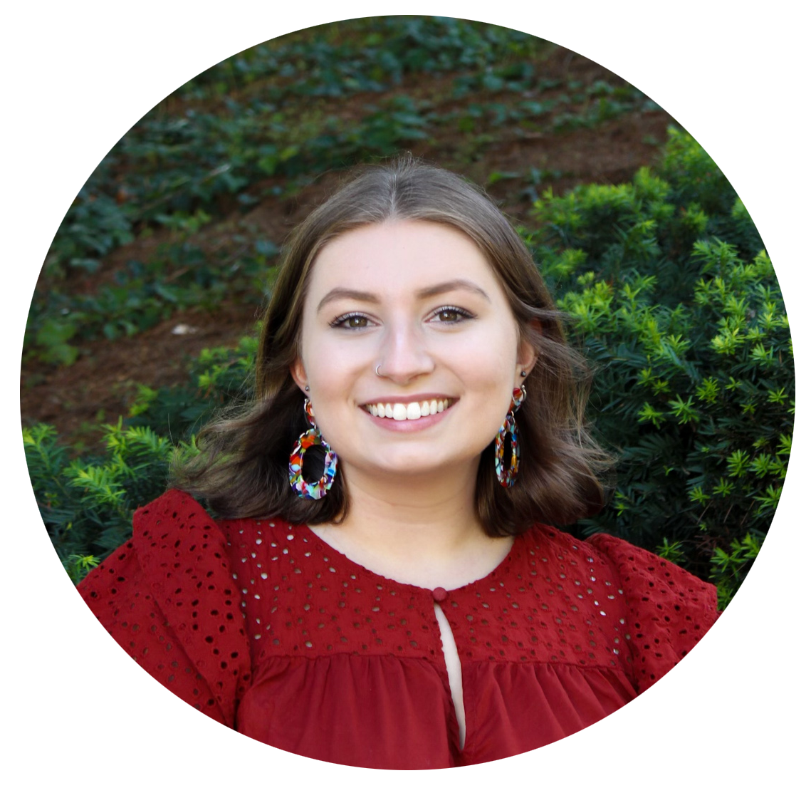 Maggie's headshot. Maggie has straight, light brown hair and hazel eyes and is smiling at the camera. She has a silver nose ring on the right side of her nose and is wearing colorful dangly earrings. She is wearing a red shirt and is surrounded by beautiful green trees and foliage.